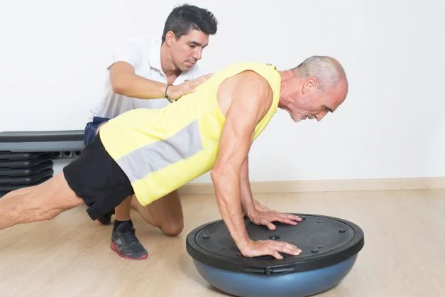 senior planking on bosu ball with personal trainer on side