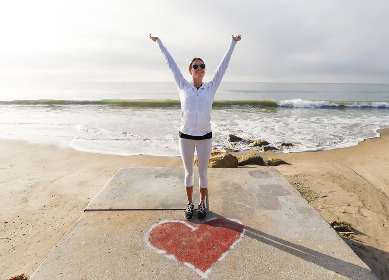 female with hands up in air on beach with drawn red heart in front of her
