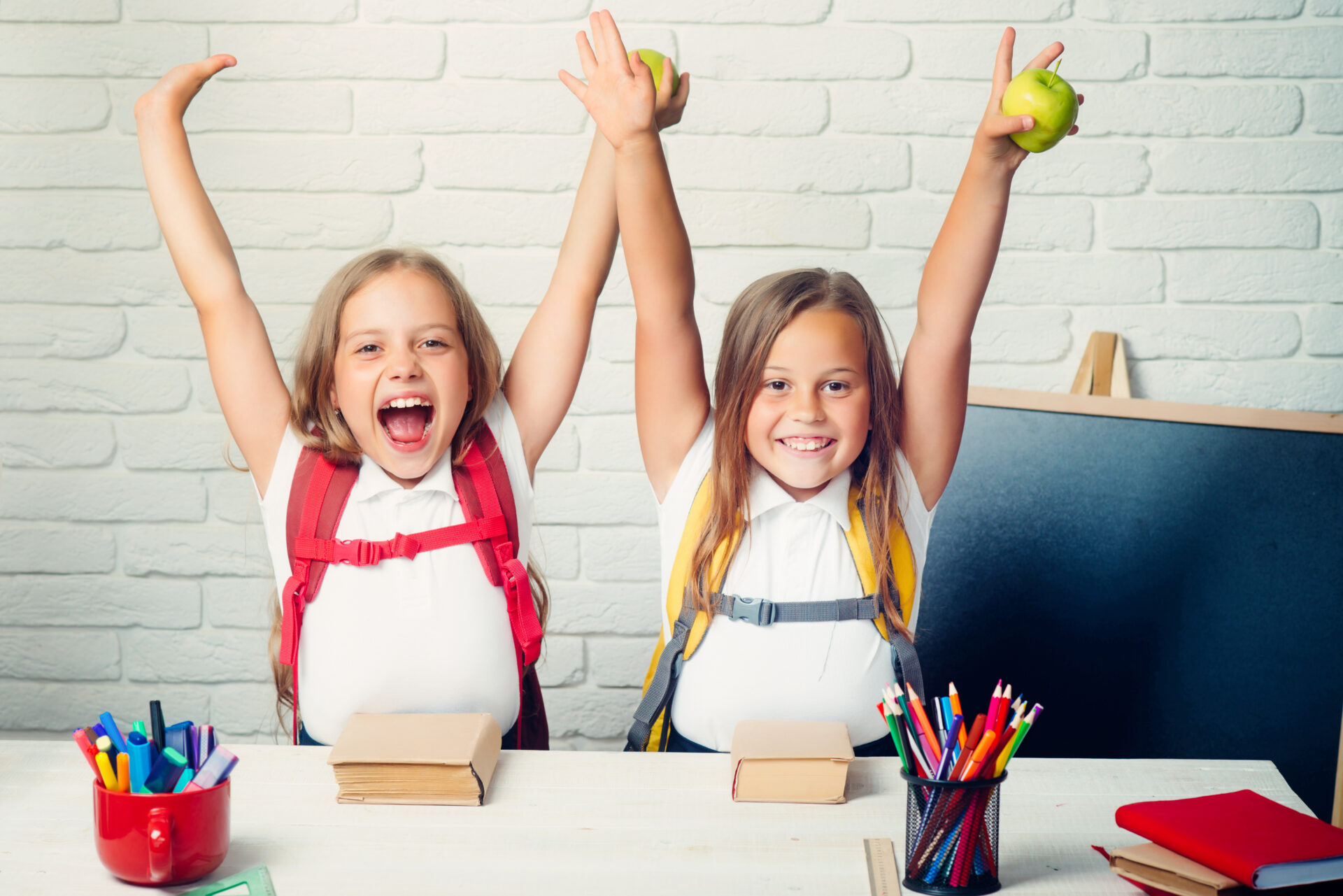 Back to school and home schooling. Happy school kids. Friendship of small pupils in classroom at knowledge day. Two happy elementary school girls with hands up in the air each holding an apple