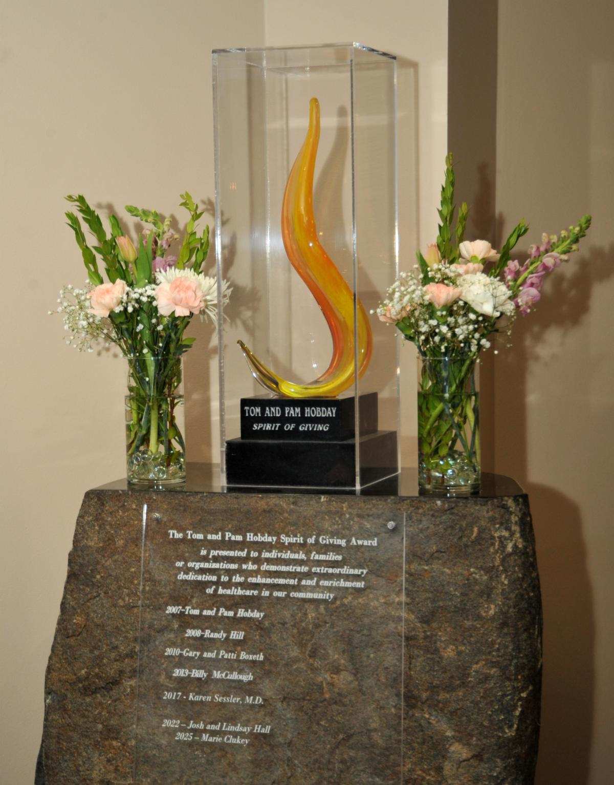 Tom and Pam Hobday Spirit of Giving Award displayed in case surrounded by bouquets of flowers