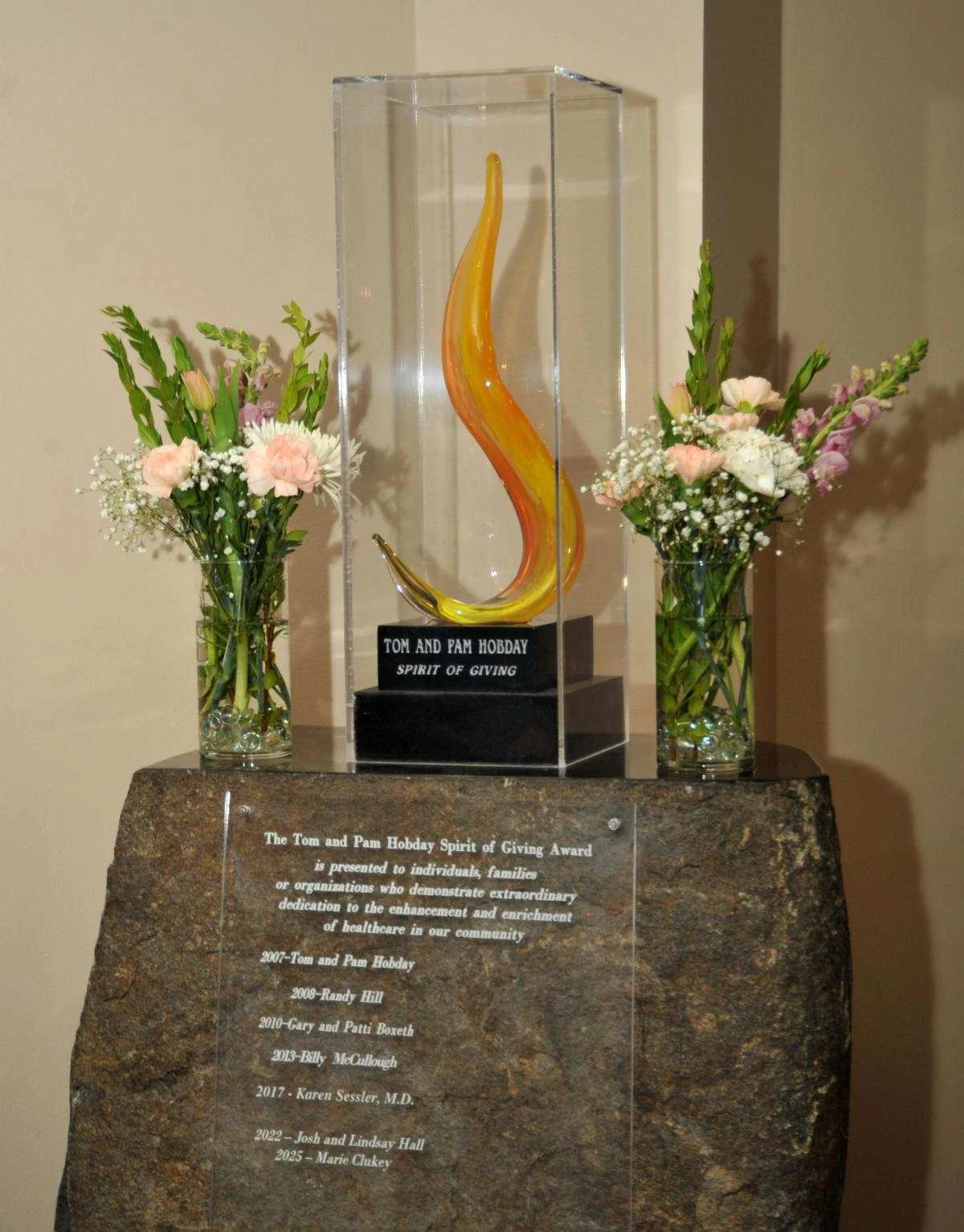Tom and Pam Hobday Spirit of Giving Award displayed in case surrounded by bouquets of flowers