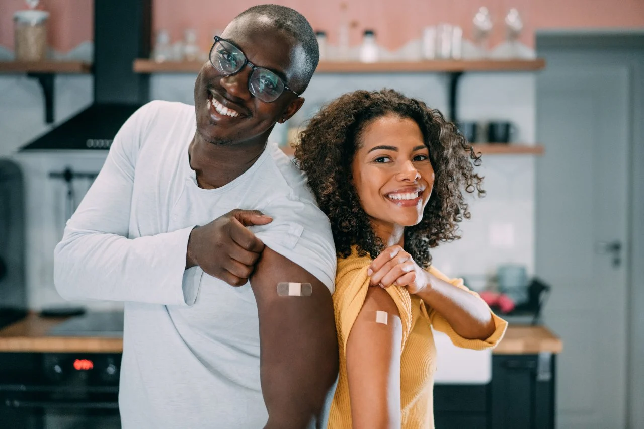 2 adults stand back to back showing bandaids on their upper arms